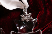 A close-up of a clear faceted star-shaped ornament with a silver snowflake charm engraved with '2026', hanging from a white ribbon against a dark red velvet background.