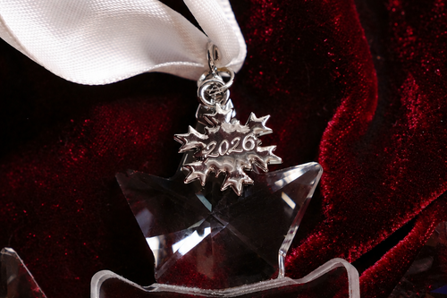 A close-up of a clear faceted star-shaped ornament with a silver snowflake charm engraved with '2026', hanging from a white ribbon against a dark red velvet background.